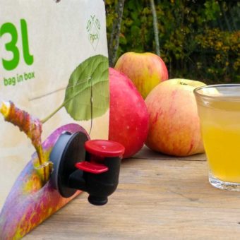 Eine Streuobstsaftbox auf einem Gartentisch mit 3 Äpfeln der Sorte Wiltshire und einem vollen Glas Streuobsaft im Bauerngarten des Bienenhof Hallertau der Imkermei Oswald.