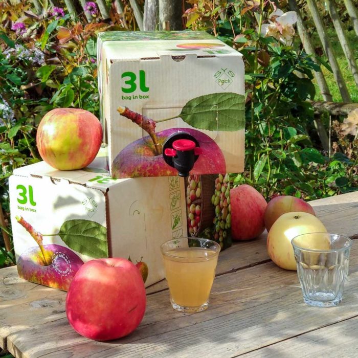 Ein Tisch im Bauerngarten, auf dem zwei Kartons bag-in-box Streuobst-Direktsaft stehen, zusammen mit einem leeren und einem vollen Glas Streuobst-Bio-Apfel-Birnen-Quitten-Saft, une eineigen Äpfeln der Sorte Korbinian und Wiltshire. Foto: Imkerei Oswald bio-honig.com im November 2023.