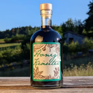 Flasche Honig-Kräuterlikör von Imkerei Oswald, handwerklich hergestellt mit echtem Bienenhonig. Die dunkle Kräuterspirituose in der Glasflasche mit Holzstopfen zeigt ein Etikett mit "Honig Kräuter" und Illustrationen von Sternanis und Waben. Im Hintergrund ist ein Bienenstand auf einer Wiese unter blauem Himmel zu sehen.