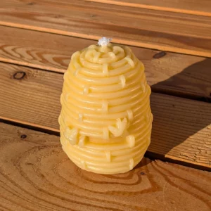 Detaillierte, hellgelbe Bienenkorbkerze hell oval von Imkerei Oswald aus echtem Bienenwachs mit kleinen Bienenmodellen am Flugloch und Baumwolldocht auf Holztisch. Sie bilden einen Flechtkorb oder Strohbienenkorb ab, der traditonell für die Haltung von Bienen verwendung fand.