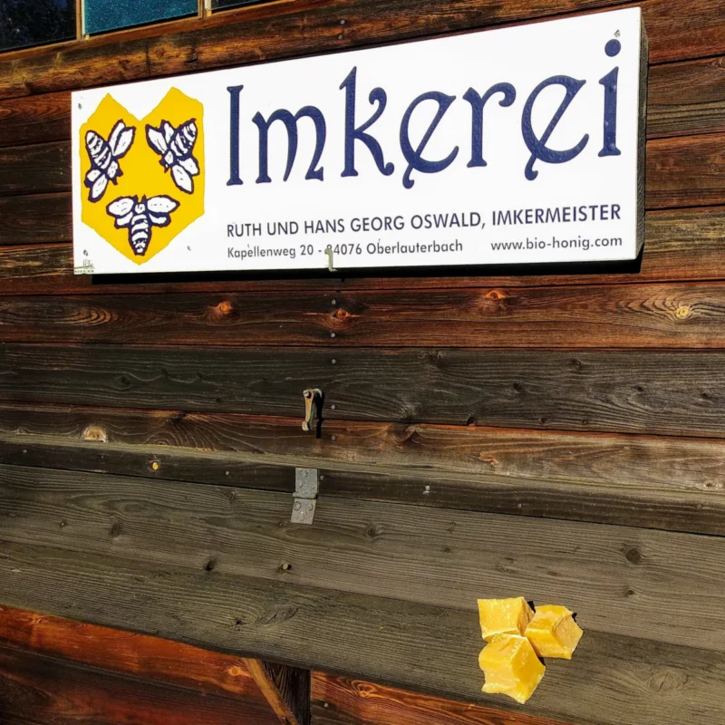 Das erste Imkerei-Schild der Imkerei Oswald von ca. 1999 an der Außenwand des Bienenhauses von 1946. Auf dem Anflugbrett mit geschlossenen Flugklappen liegen drei Stücke Bienenwachs aus der Kerzenherstellung. Foto: Imkerei Oswald.
