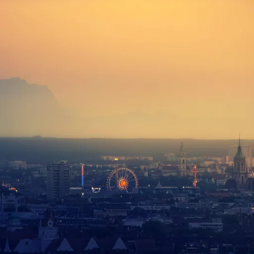 Alt-Text (für Barrierefreiheit & SEO): Münchner Skyline in der warmen Abenddämmerung mit dem beleuchteten Riesenrad auf der Theresienwiese und der schemenhaften Silhouette der Alpen am fönigen Horizont.