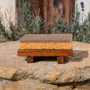 Eine rustikale Seifenablage aus Holz auf kleinen Beinen hält einen gelb-braunen Schwamm, präsentiert im Freien auf einem Naturstein vor Lavendelpflanzen.